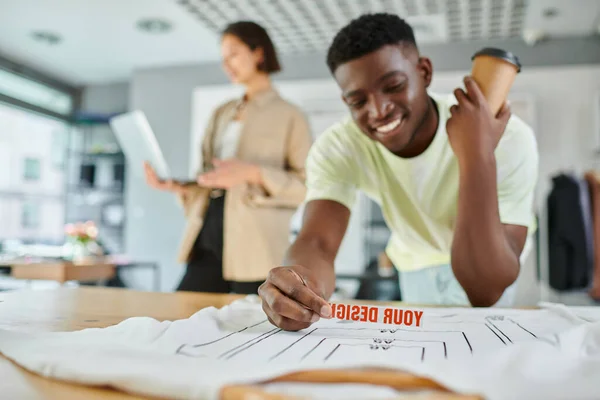 Happy african american designer with text template near clothes and asian colleague with laptop — Stock Photo