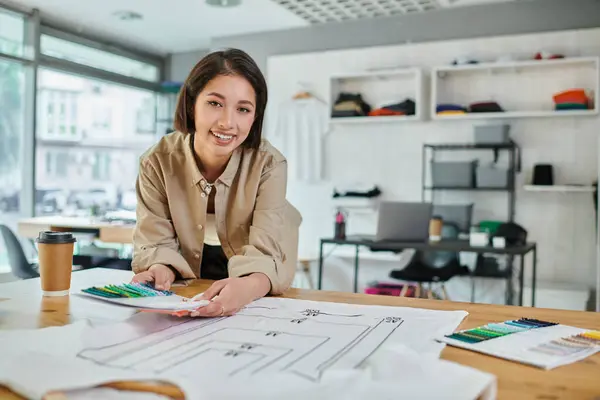 Freudige kreative asiatische Designerin mit Farbmustern in der Nähe von Kleidung mit Formatgrößen im Druckstudio — Stockfoto