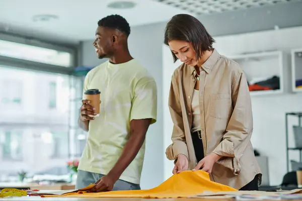 African american man with coffee to go near asian designer working with fabric in print studio — Stock Photo