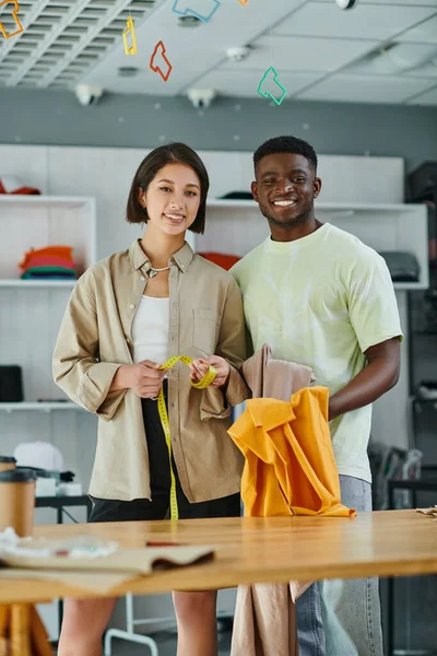Happy interracial clothes designers with fabric and measuring tape looking at camera in print studio — Stock Photo