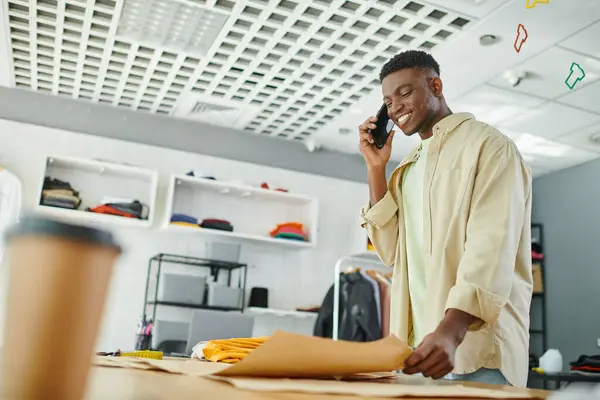 Felice stilista afroamericano che parla sul telefono cellulare vicino a modelli di cucito in studio di stampa — Foto stock
