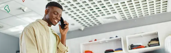 Alegre africano americano diseñador de moda hablando en el teléfono móvil en moderno estudio de impresión, pancarta - foto de stock