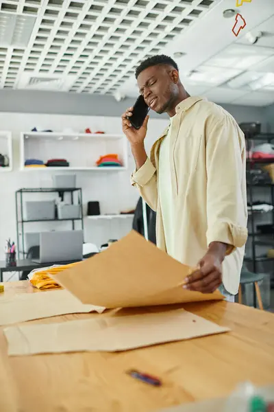 Вдумливий афроамериканський дизайнер моди розмовляє на мобільному телефоні біля швейних візерунків в ательє — стокове фото