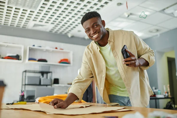 Designer americano africano de sucesso com smartphone sorrindo para a câmera perto de padrões de costura — Fotografia de Stock