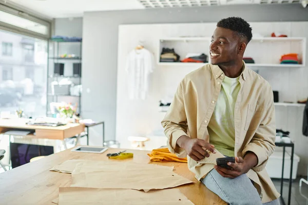 Joyful african american entrepreneur sitting with smartphone and looking away near sewing patterns — Stock Photo