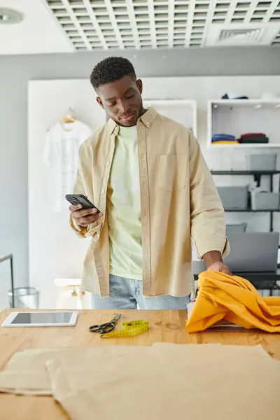 Designer americano africano com smartphone tocando amostras de tecido perto de tesoura e tablet digital — Fotografia de Stock
