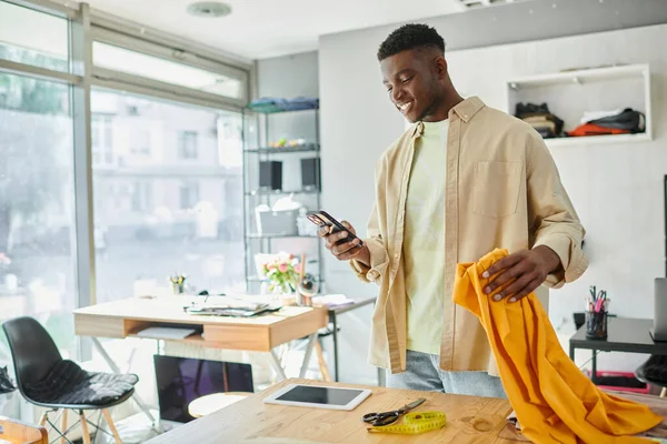 Усміхнений афроамериканський дизайнер дивиться на смартфон, тримаючи зразки тканини в друкованій студії — стокове фото