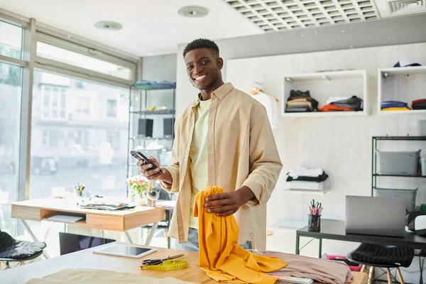 Designer americano africano feliz com amostras de smartphone e tecido olhando para a câmera no estúdio de impressão — Fotografia de Stock