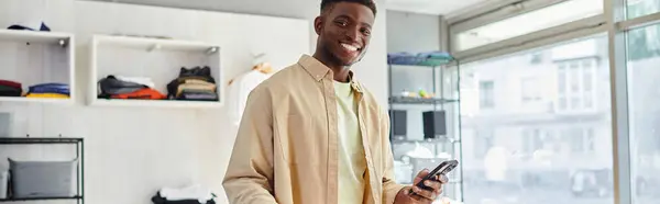 Designer de moda afro-americano satisfeito com smartphone olhando para a câmera no estúdio de impressão, banner — Fotografia de Stock