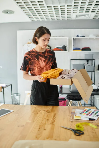 Stilista di moda asiatica elegante disimballando i vestiti dalla scatola di cartone vicino ai campioni di colore in studio di stampa — Foto stock