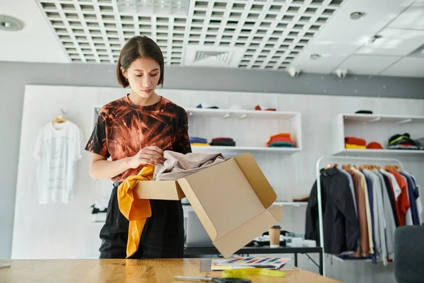 Молодий азіатський дизайнер моди розпаковує одяг з картонної коробки біля кольорової палітри в друкованій студії — стокове фото