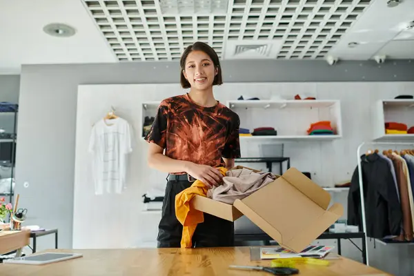 Alegre asiático empreendedor segurando caixa de papelão com roupas e olhando para a câmera no atelier impresso — Fotografia de Stock