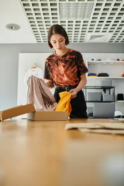 Stilvolle asiatische Modedesignerin packt Karton mit Kleidungsstücken aus, während sie im Druckstudio arbeitet — Stockfoto