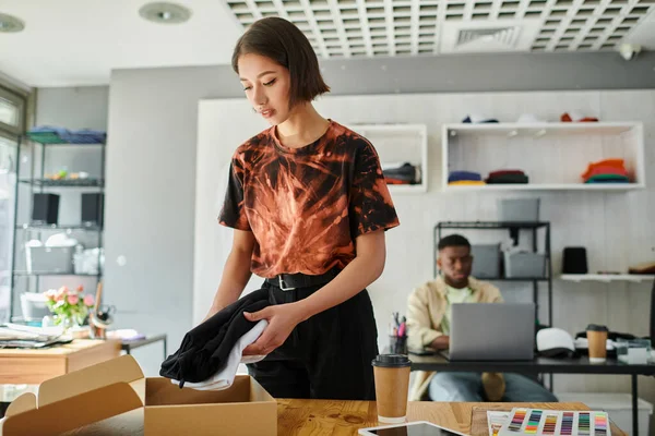 Asiática mulher embalagem roupas em caixa de papelão perto de Africano americano designer no laptop no estúdio de impressão — Fotografia de Stock