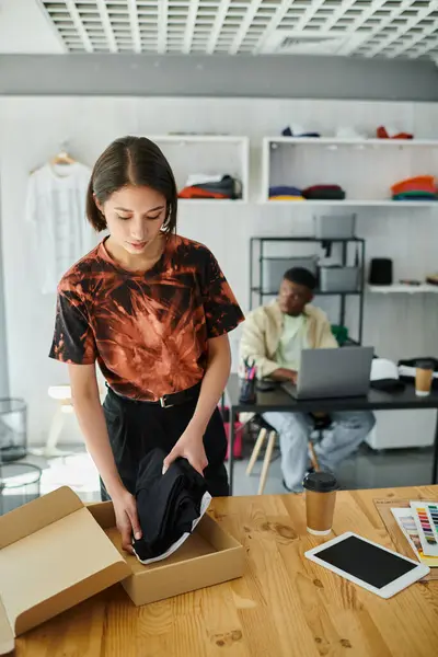 Азиатка упаковывает одежду в коробку рядом с африканским американским дизайнером на ноутбуке в печатном ателье — стоковое фото