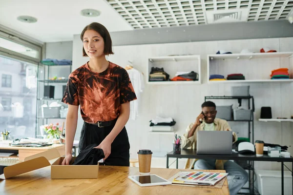 Happy asian woman packing clothes and near african american colleague at laptop in print studio — Stock Photo