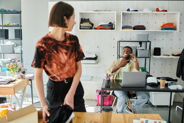 Азиатка упаковывает одежду рядом с веселым африканским американским коллегой разговаривающим по смартфону — стоковое фото
