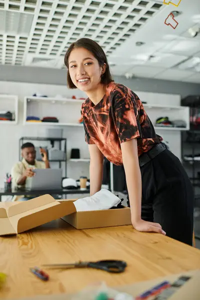 Alegre asiático mulher olhando para câmera perto caixa de papelão e africano americano colega no print studio — Fotografia de Stock