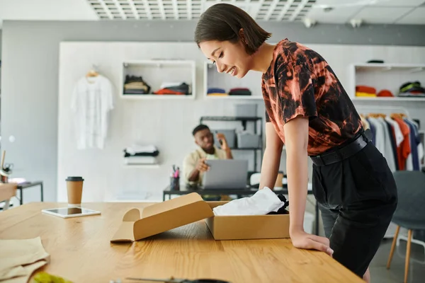 Happy asian designer near box with garments and african american colleague working in print studio — Stock Photo
