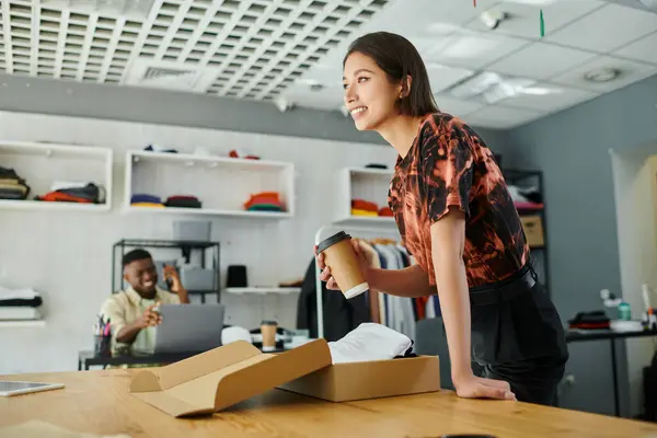 Pleased asian woman with coffee to go near clothes in carton box and african american man colleague — Stock Photo