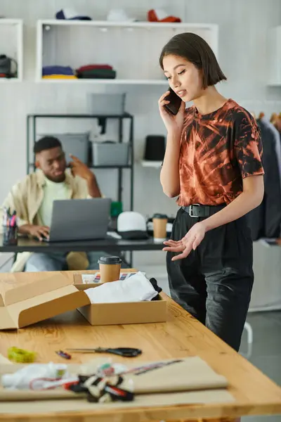 Asian designer talking on smartphone near clothes in carton box and african american man at laptop — Stock Photo