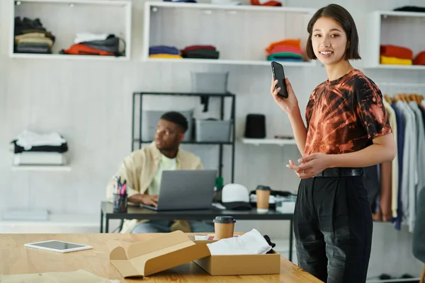 Asian woman with smartphone smiling near clothes in carton box and african american man at laptop — Stock Photo