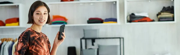 Alegre asiático designer de moda com telefone móvel olhando para a câmera no estúdio de impressão têxtil, banner — Fotografia de Stock