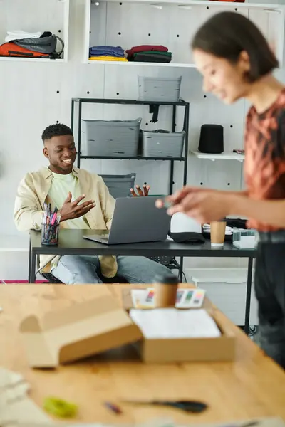 Happy african american designer having video chat on laptop near asian woman taking photo of clothes — Stock Photo