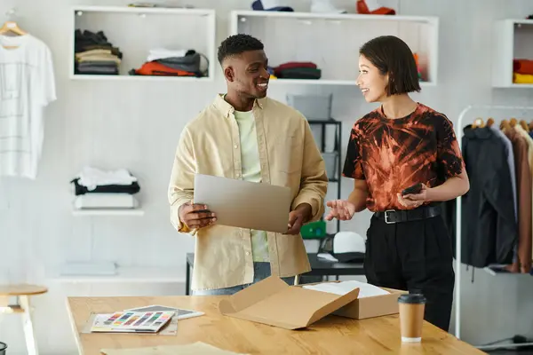 Designers inter-raciais felizes com gadgets falando perto da caixa de papelão com roupas no estúdio de impressão — Fotografia de Stock