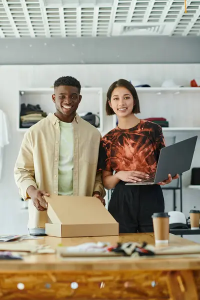 Emprendedores multiculturales alegres con portátil y caja de cartón mirando a la cámara en el estudio de impresión - foto de stock