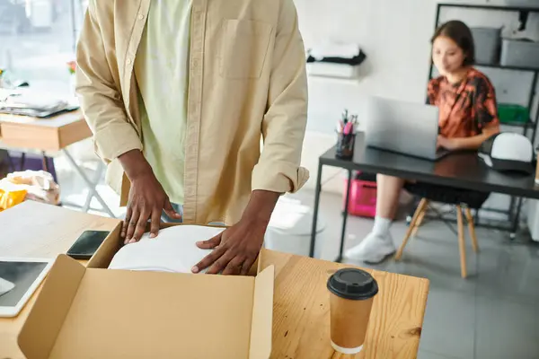 African american man packing clothes in carton box near asian designer at laptop in print studio — Stock Photo