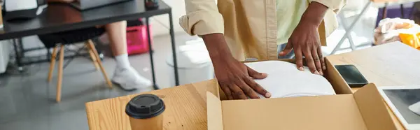 Vista ritagliata dell'uomo africano americano che imballa i vestiti in scatola di cartone vicino alla tazza di carta in atelier, banner — Foto stock