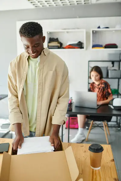 Piacere africano americano uomo imballaggio vestiti in scatola di cartone vicino asiatico designer a laptop in atelier — Foto stock