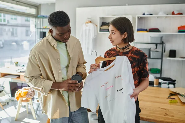 Asiático diseñador mostrando moda camiseta con impresión a africano americano colegas con café a ir - foto de stock