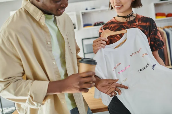 Vue recadrée de designers multiethniques souriants avec café à emporter et t-shirt élégant en studio d'impression — Photo de stock