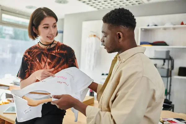 Усміхнена азіатська жінка вказує на текст, надрукований на футболці біля афроамериканського дизайнера в ательє — стокове фото