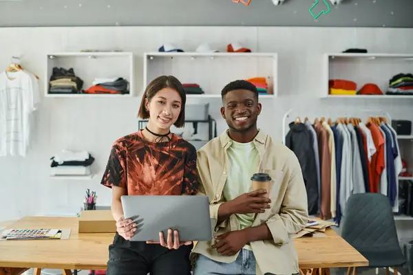 Designers multiétnicos alegres com laptop e café para ir olhando para a câmera no estúdio de impressão têxtil — Fotografia de Stock