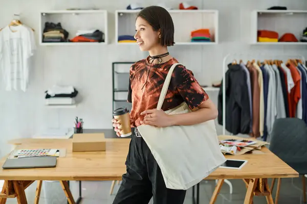 Радостная и стильная азиатская женщина с пакетом для покупок и напитком на вынос в современной типографии — стоковое фото