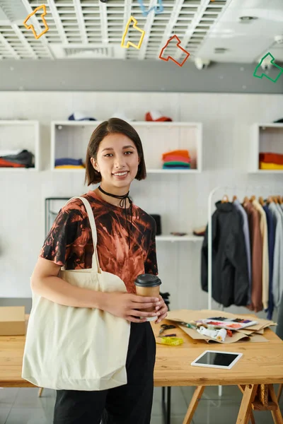 Happy asian designer with canvas shopping bag and coffee to go looking at camera in print studio — Stock Photo