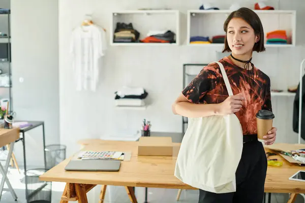 Задоволений азіатський дизайнер з полотном сумка для покупок і паперовий стаканчик, дивлячись далеко в друкованій студії — стокове фото