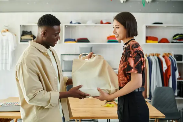 Smiling asian designer giving canvas shopping bag to african american colleague in print studio — Stock Photo