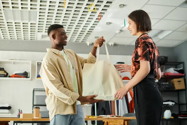 Feliz Africano americano designer segurando lona saco de compras perto asiático colega na oficina de impressão — Fotografia de Stock
