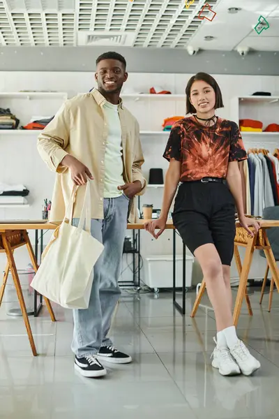 Cheerful multiethnic fashion designers with canvas shopping bag looking at camera in print studio — Stock Photo
