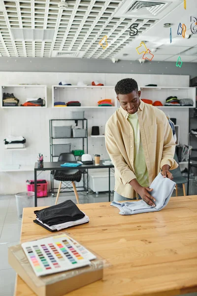 Fröhlicher afrikanisch-amerikanischer Mann faltet Kleidung auf dem Tisch in einem modernen Druckereistudio, Kleinunternehmen — Stockfoto