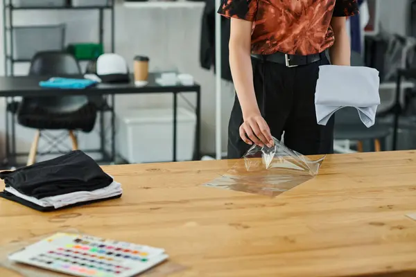 Обрізаний вид молодої жінки, що упаковує складений одяг у поліетиленовий пакет на столі в друкарні — стокове фото