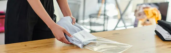 Обрізаний вид жіночої працівниці упаковки складеного одягу в поліетиленовий пакет на столі в друкованій студії, банер — стокове фото
