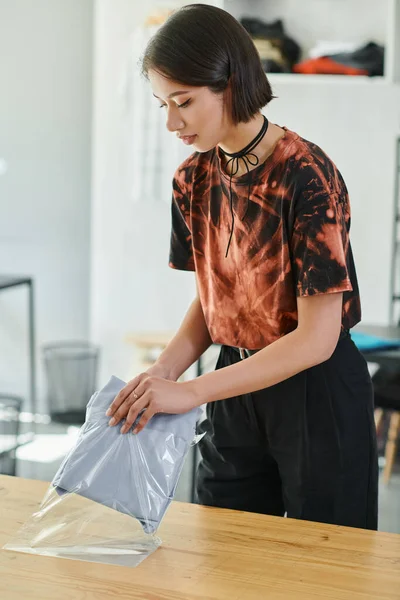 Mujer asiática diligente embalaje cuidadosamente doblado prendas en bolsa de plástico en atelier de impresión, pequeña empresa - foto de stock