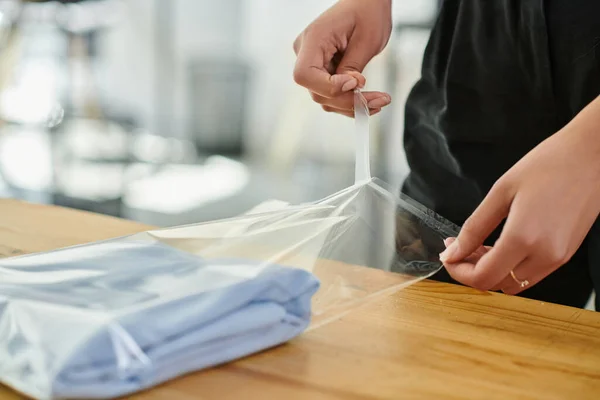 Vista parcial de trabajadora cualificada empacando ropa doblada en bolsa de plástico en la mesa en el estudio de impresión - foto de stock