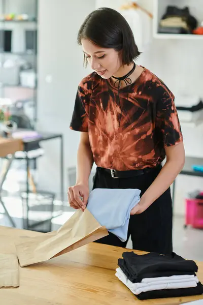Joven y elegante mujer asiática embalaje doblado ropa en bolsa de papel en estudio de impresión, pequeña empresa - foto de stock