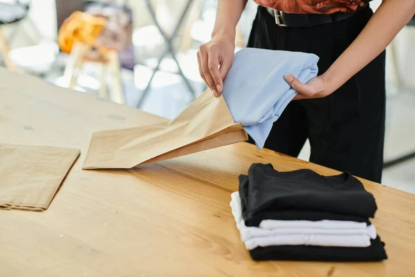 Vista recortada de mujer calificada embalaje doblado ropa en bolsa de papel en el estudio de impresión, pequeña empresa - foto de stock